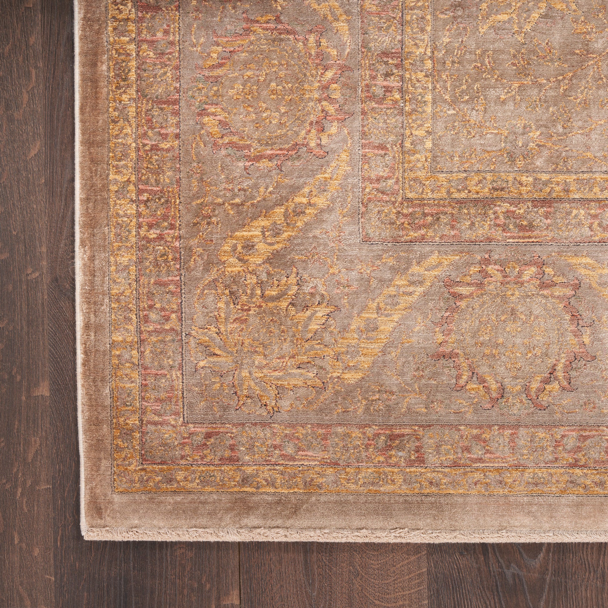 Beige Persian-style rug with gold, taupe and rust patterns and ornate border, fringe edge on dark wooden floor.