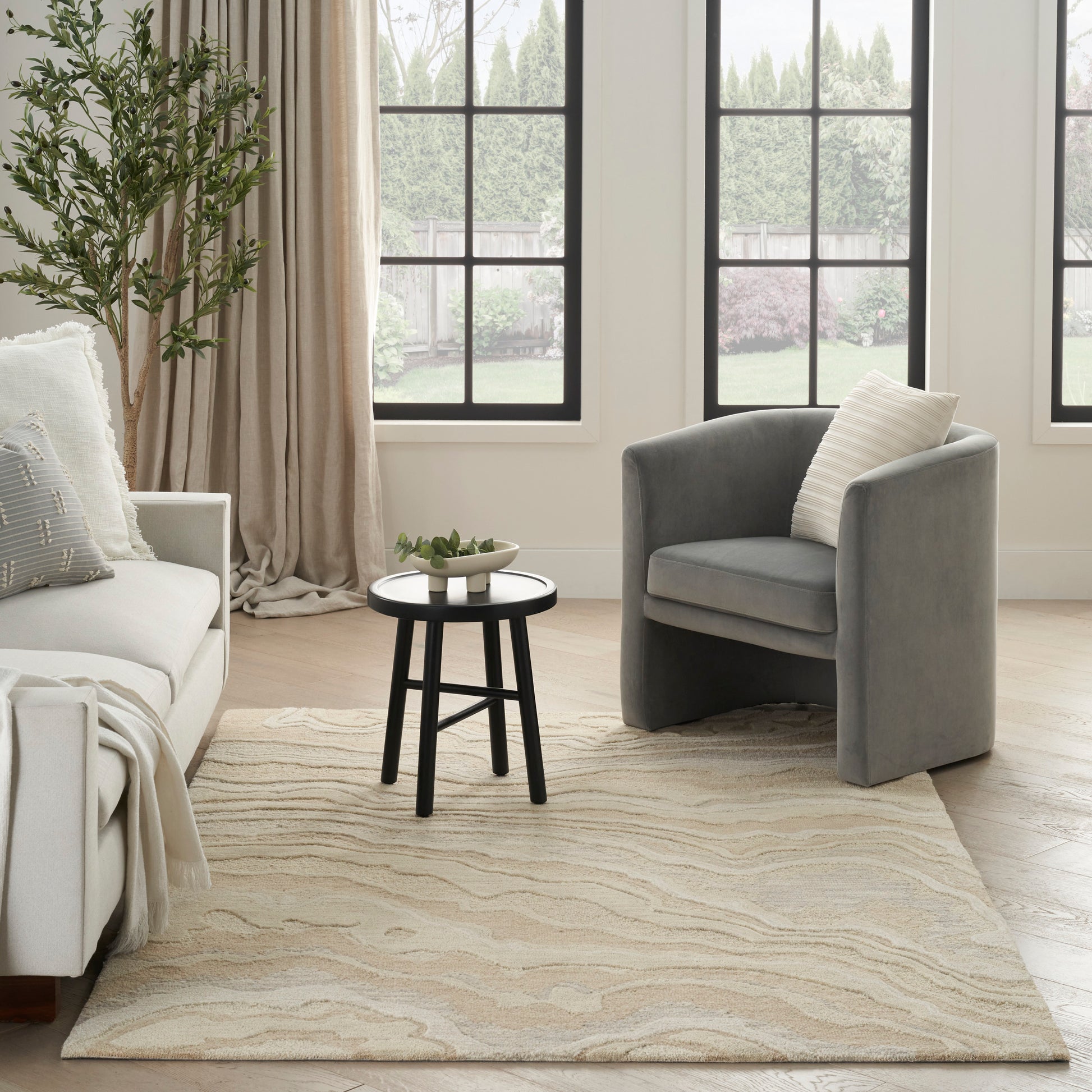 Bright living room with beige textured area rug, gray curved armchair, white sofa, black round table, and greenery.