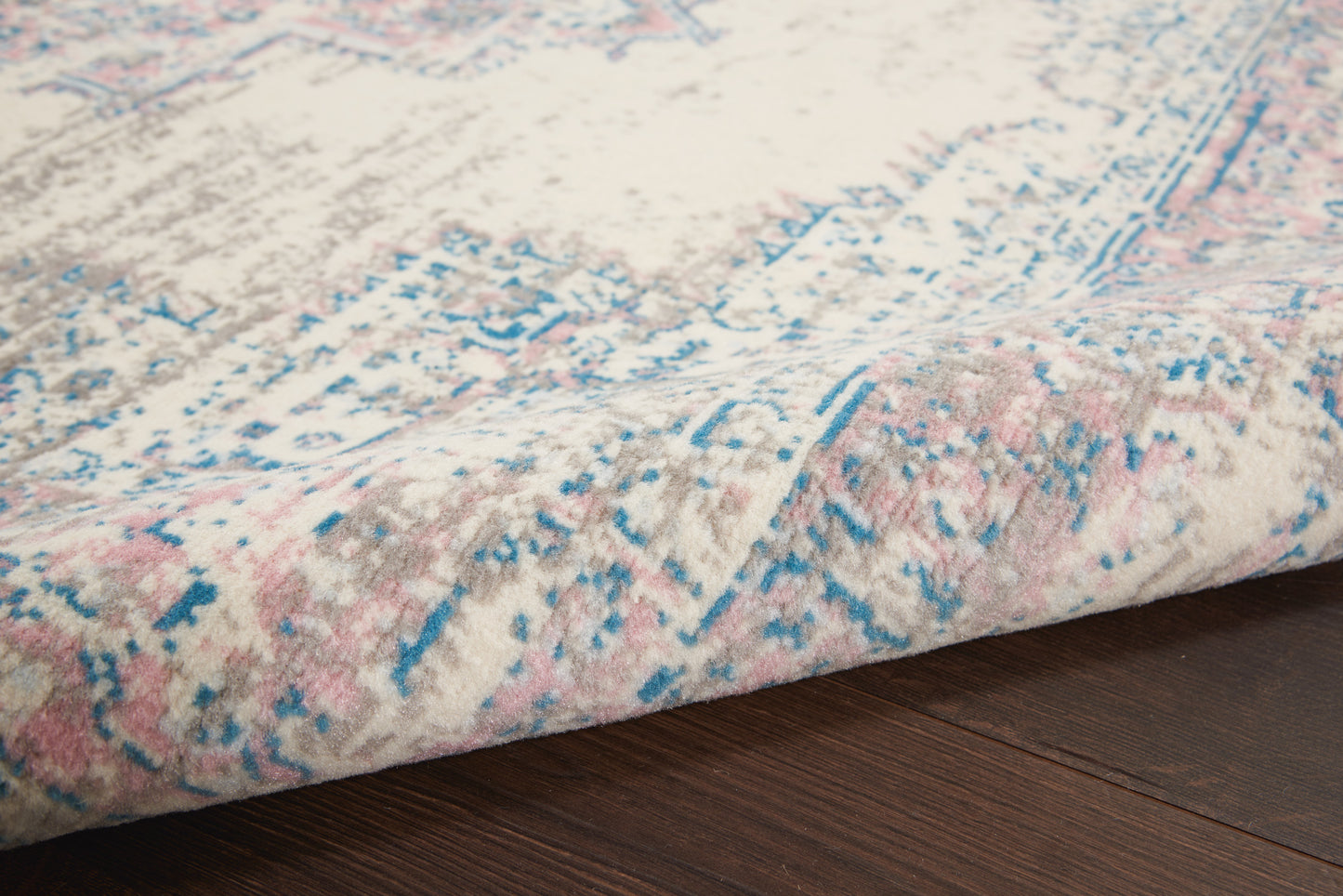 Pastel area rug with pink, blue and gray distressed pattern; folded edge on dark wood floor.