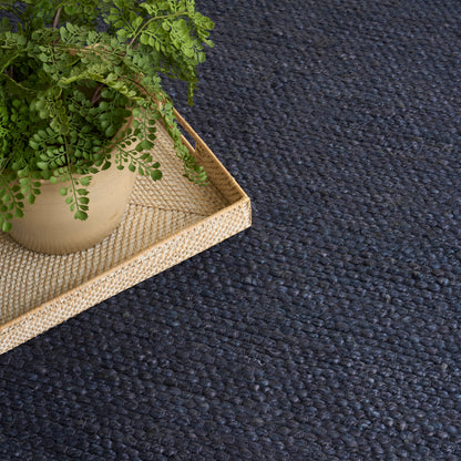 Beige ceramic pot with green fern on a light woven rattan tray atop a dark navy textured rug.