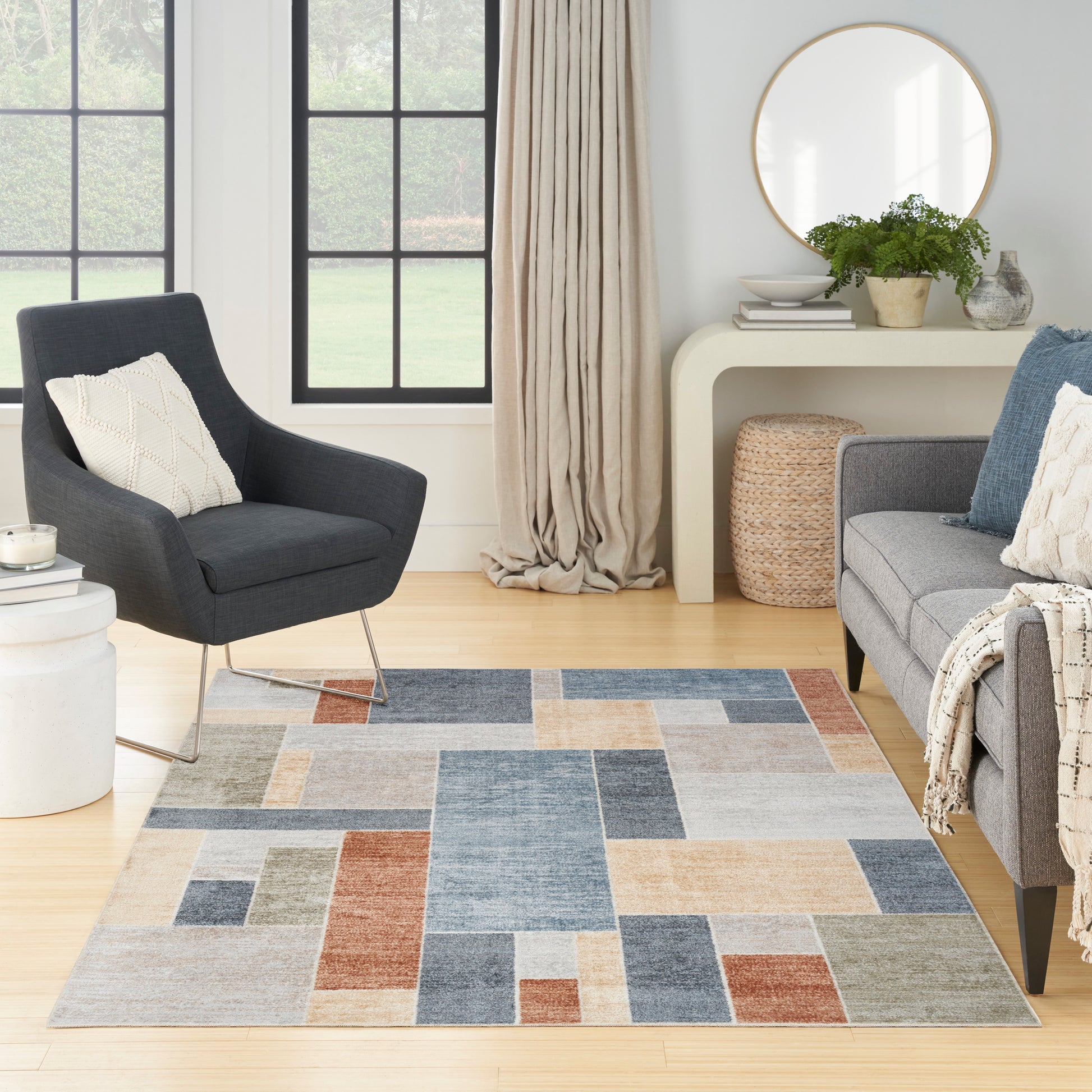 Living room with a patchwork rug in blues, beige and rust; dark gray armchair, light sofa, round gold mirror.