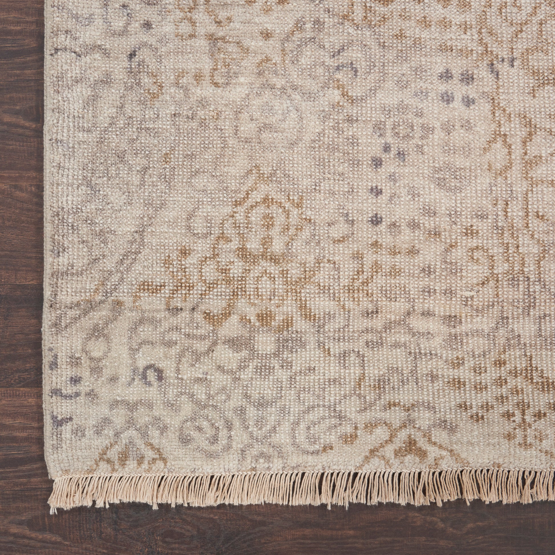 Beige traditional patterned area rug with subtle gray-brown motifs and fringe along the edge on a dark wooden floor.