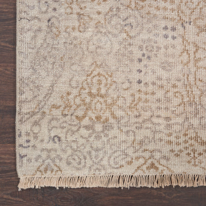 Beige traditional patterned area rug with subtle gray-brown motifs and fringe along the edge on a dark wooden floor.