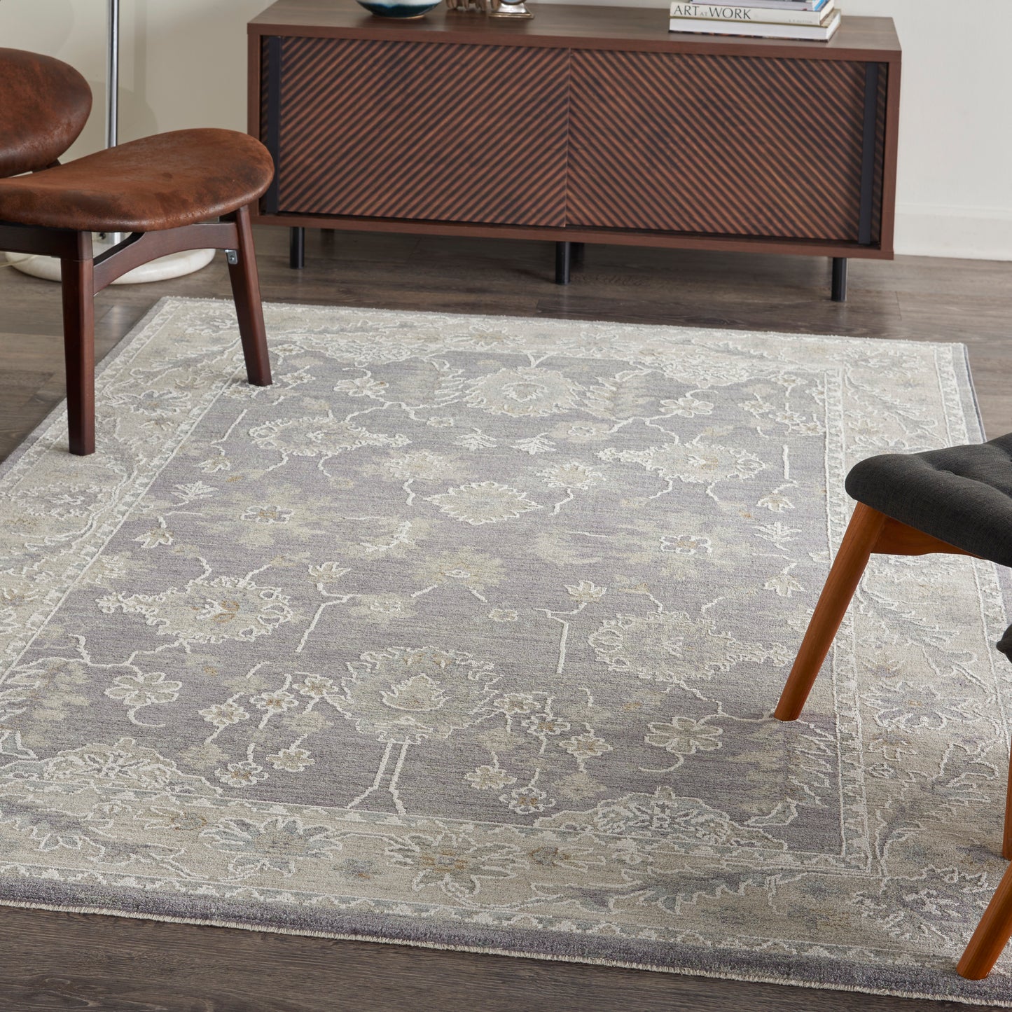 Gray floral rug with pale border under mid-century brown leather chair and dark wood chair, beside chevron sideboard.