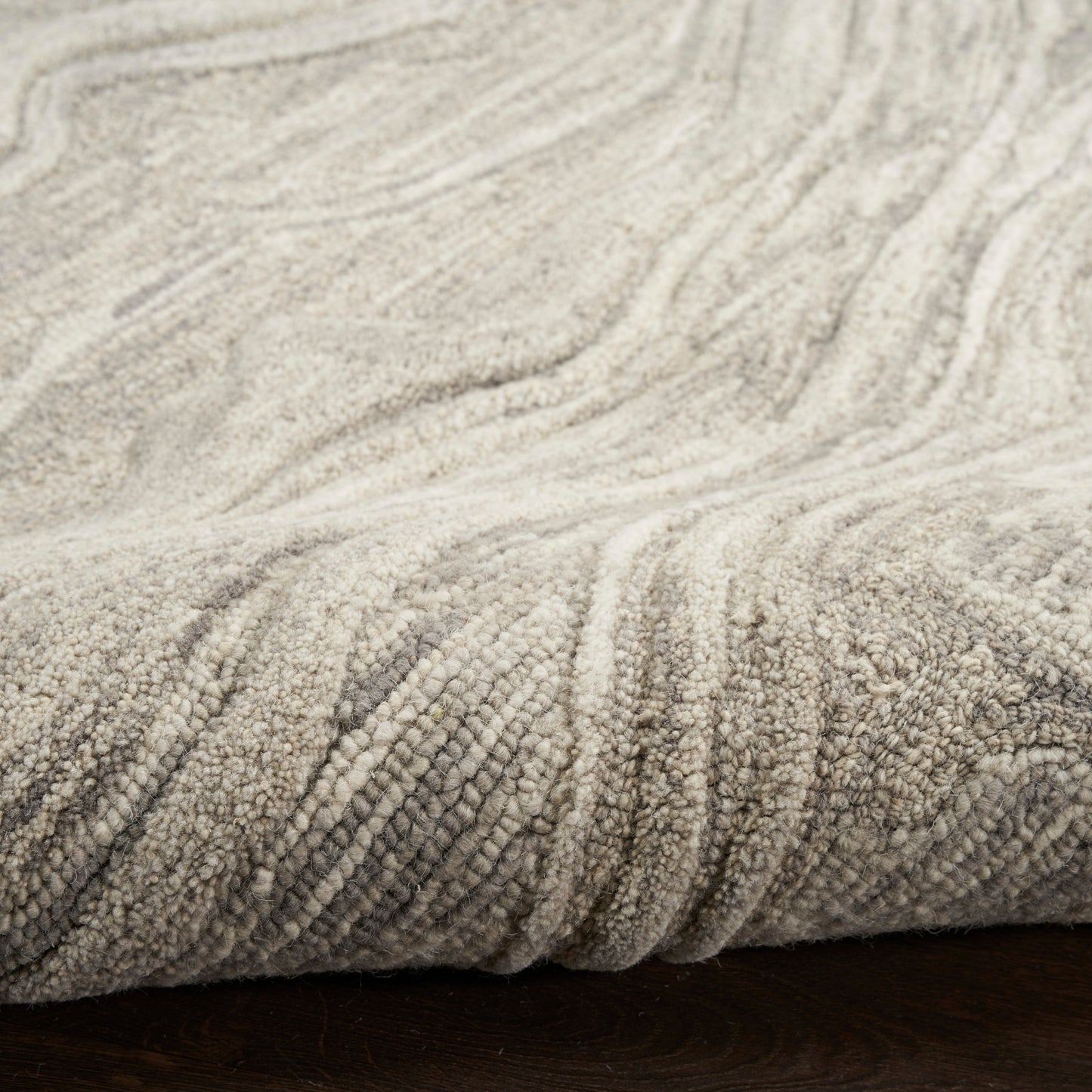 Beige textured loop-pile rug with subtle wave pattern, folded edge on dark wood floor.