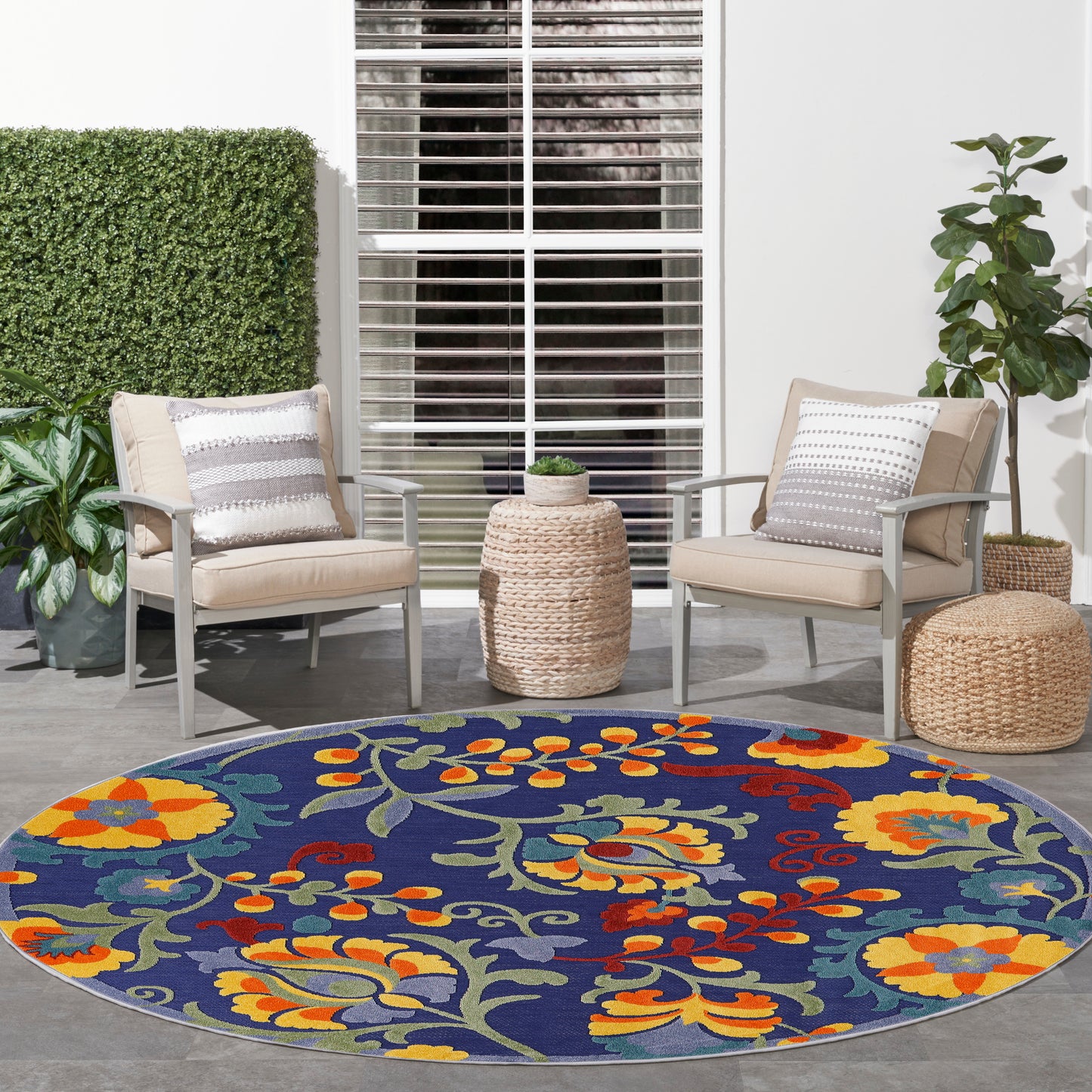 8' round navy outdoor rug with vibrant yellow, orange, red florals; two beige cushioned chairs, wicker ottomans, patio decor.