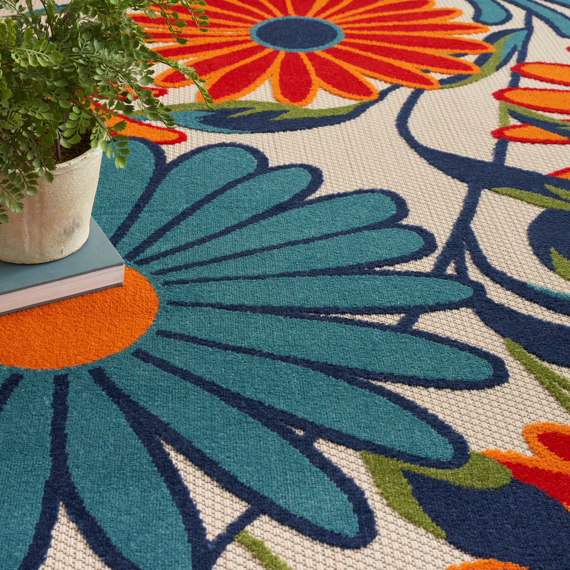 Vibrant floral area rug with turquoise petals, orange centers, navy outlines on beige background; potted plant nearby.