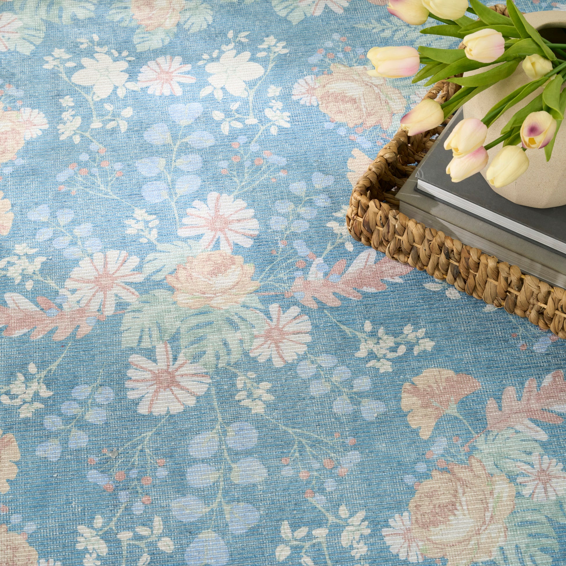 Blue floral patterned rug with pastel pink flowers and green leaves; woven basket with tulips and books resting on edge.