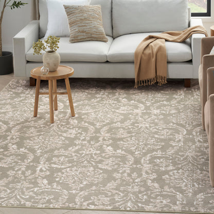 Living room with light gray sofa, beige throw blanket, patterned taupe area rug, round wooden side table with vase and plant.