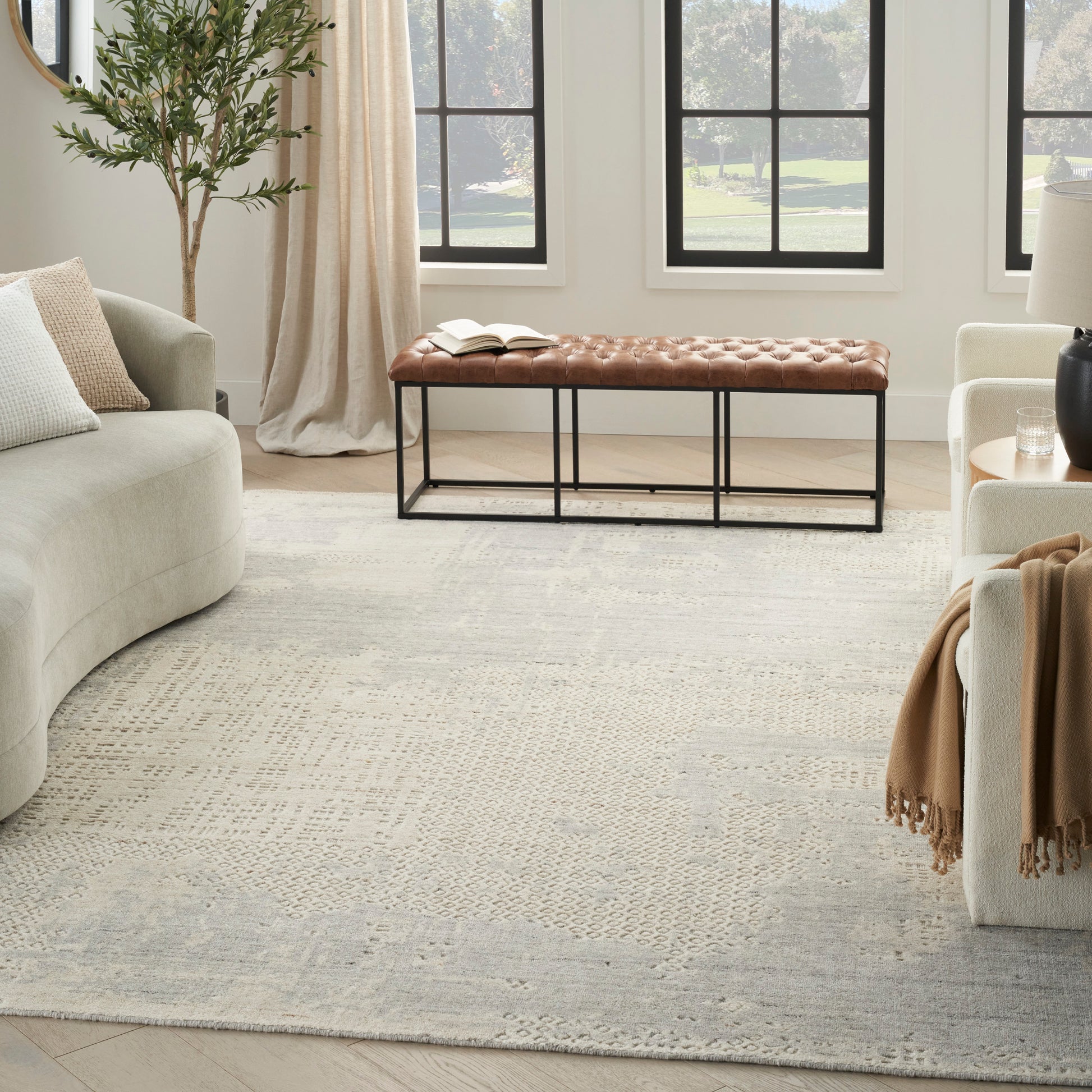 Living room with a tufted brown leather bench on a black metal frame, beige sofa, pale gray patterned rug, large windows.