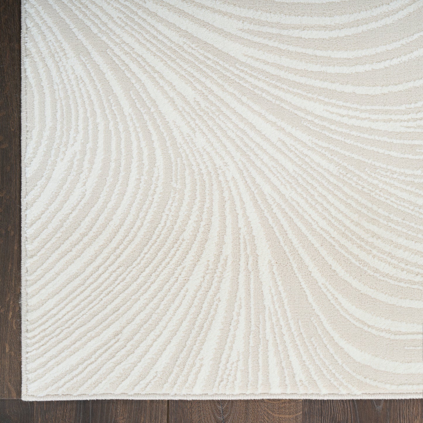 Cream textured rug with ivory curved lines in a swirling pattern, dark border along left edge, on dark wood floor.
