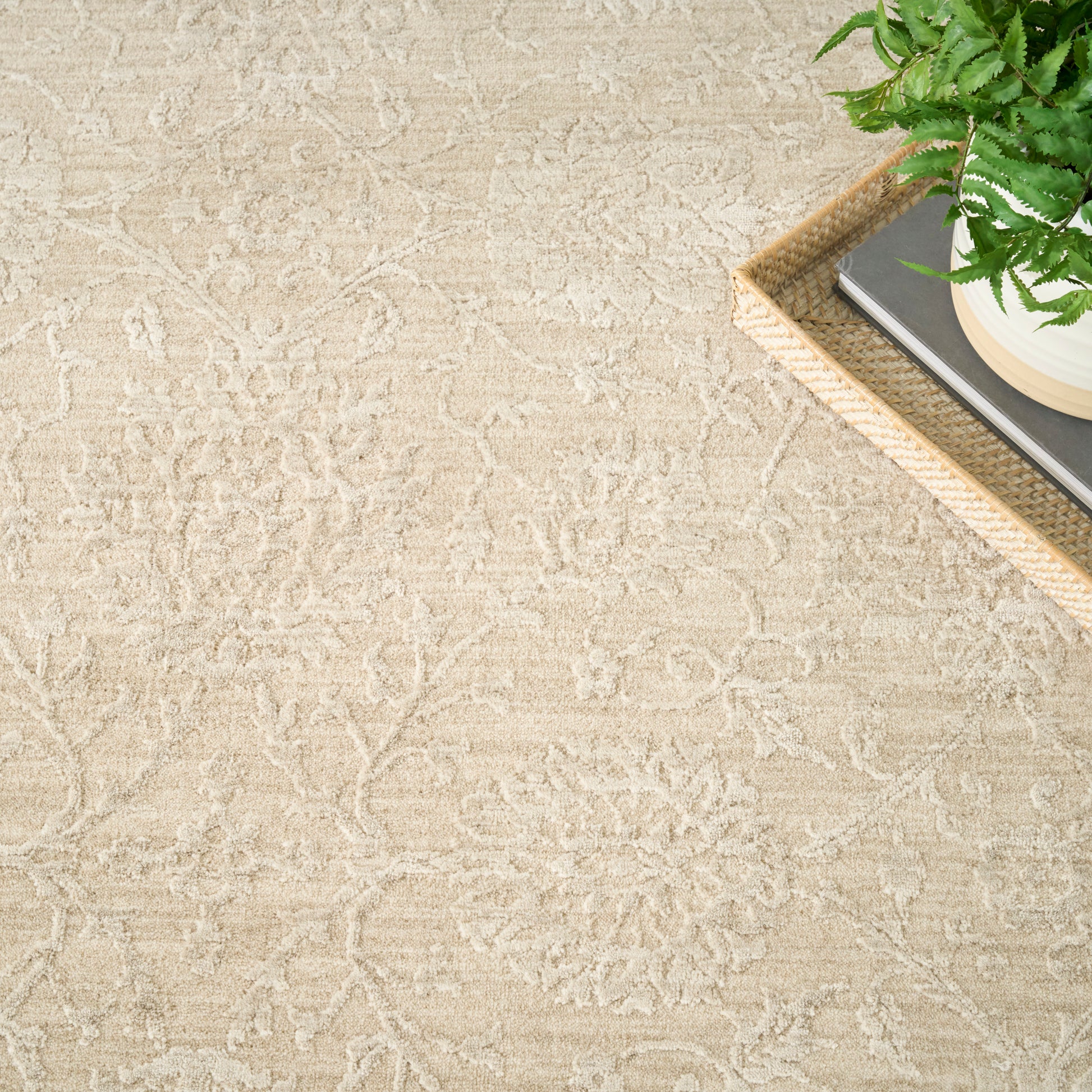 Beige patterned rug with floral motifs; corner view shows a woven tray with books & a potted fern.