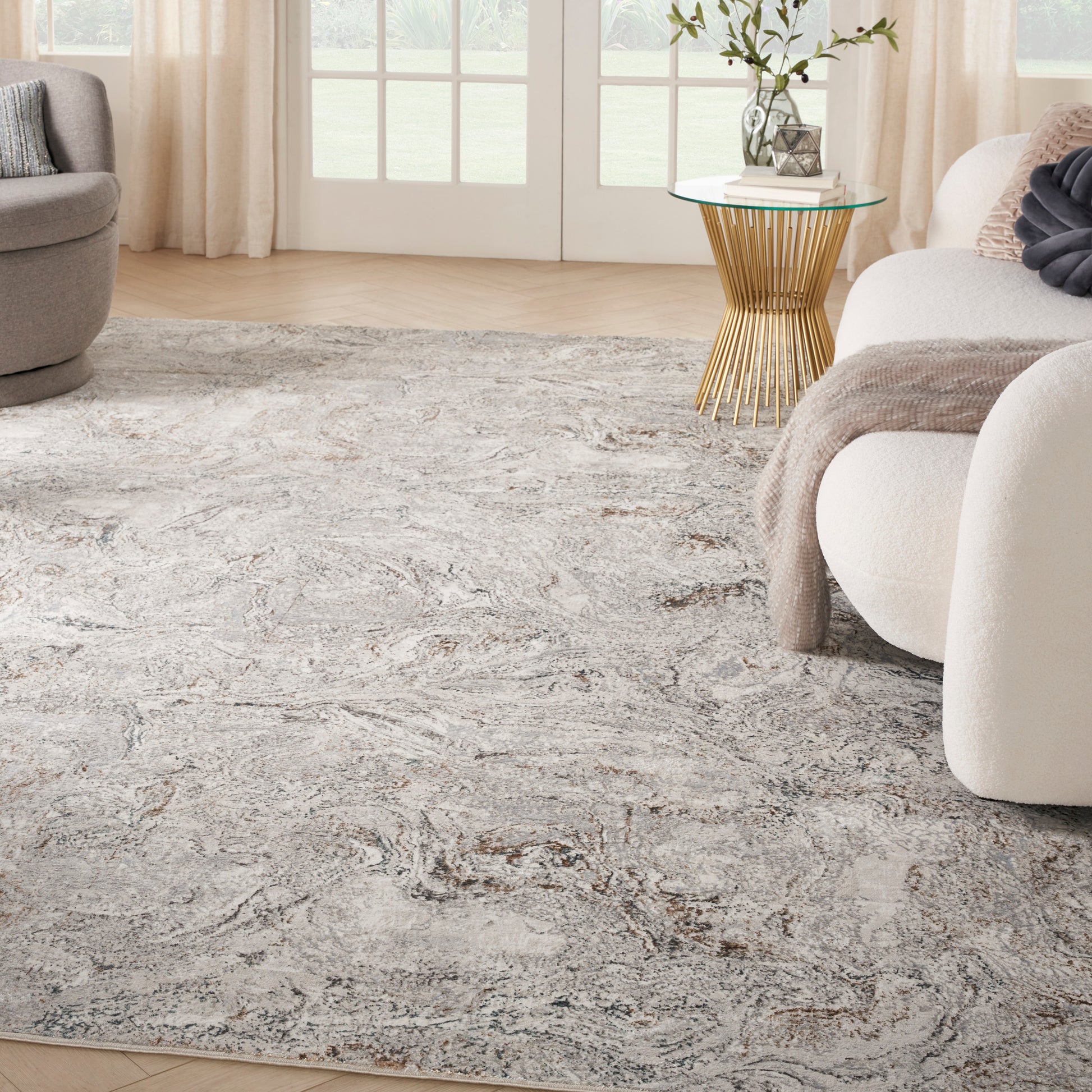 Living room with abstract gray-beige area rug, cream rounded sofa, glass-top gold pedestal table and cozy throw.