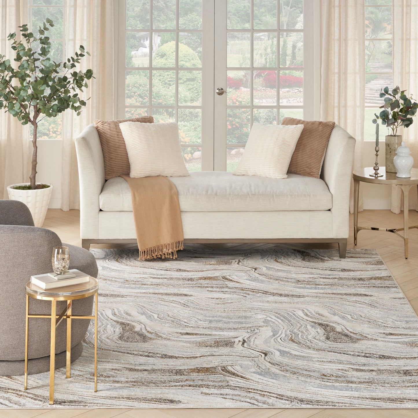 Neutral living room with cream sofa, camel throw, ivory-brown pillows, swirled gray rug, & gold glass-top side tables.