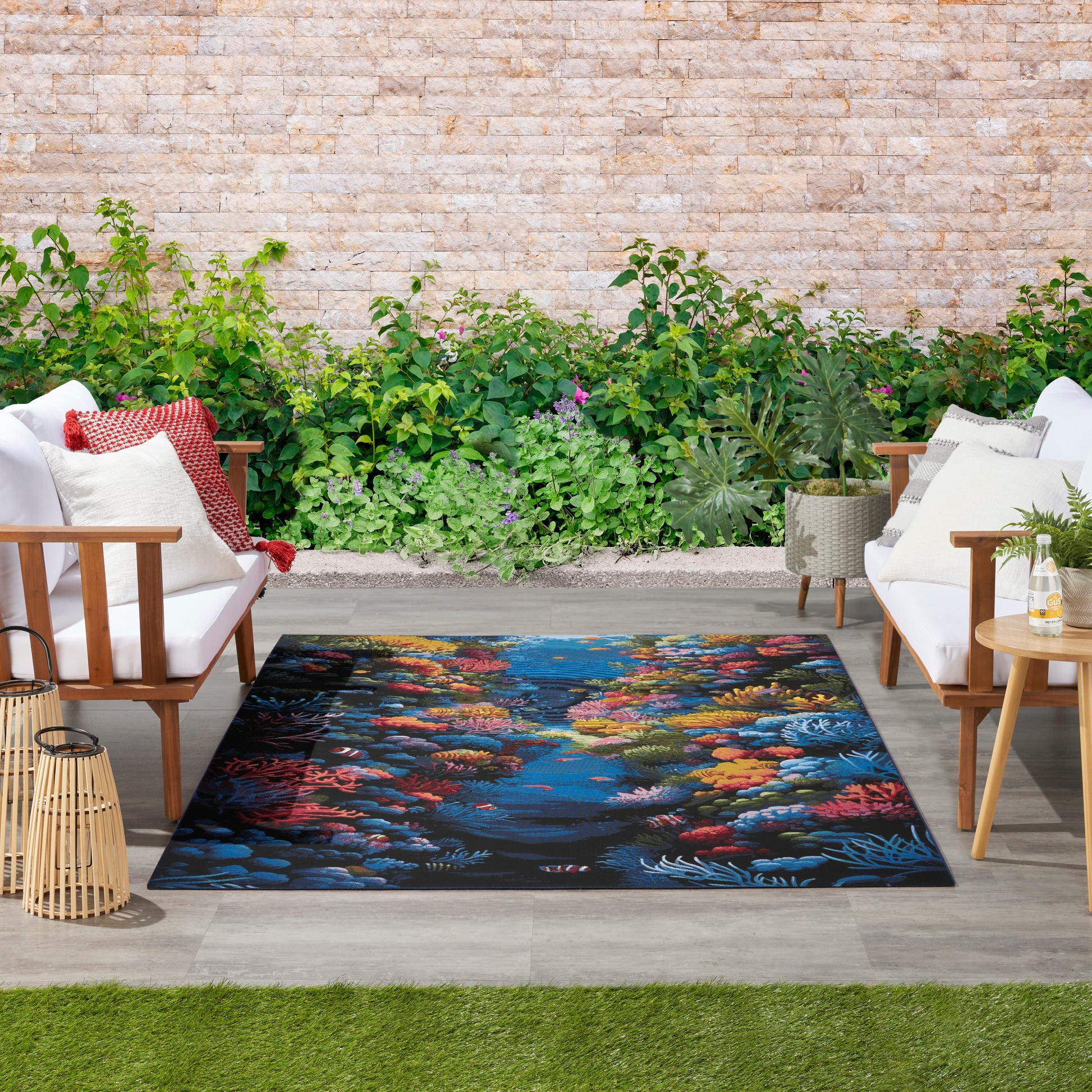 Outdoor seating area with white cushioned wooden chairs, a vibrant coral reef rug, stone wall & lush planters.