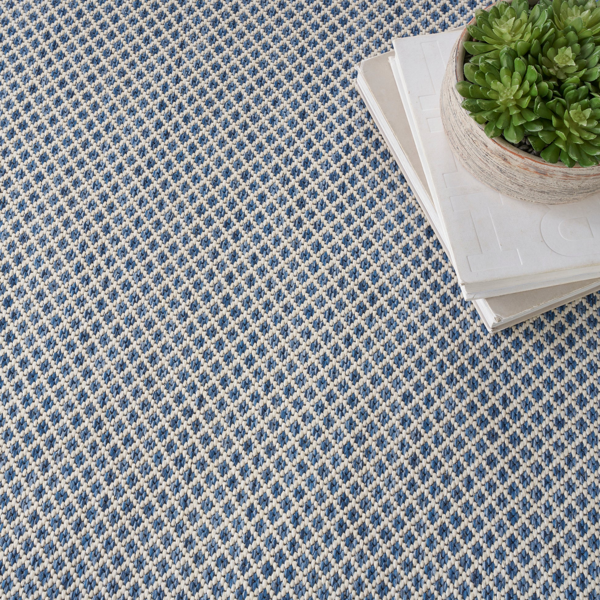 Matte gray square panel over a blue-and-white patterned rug, with a wooden edge and a potted plant in the top-right corner.