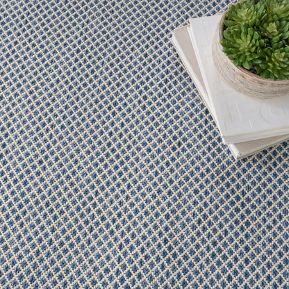 Matte gray square panel over a blue-and-white patterned rug, with a wooden edge and a potted plant in the top-right corner.