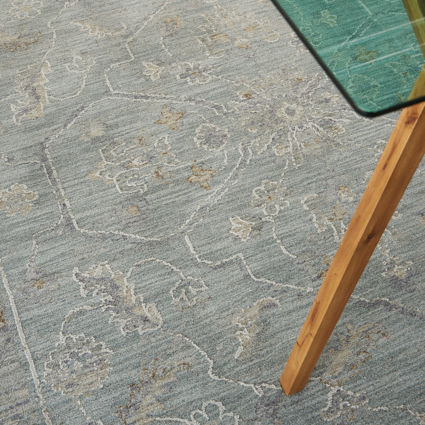 Light blue floral patterned area rug with ivory detailing; glass-topped table with a natural wood leg.