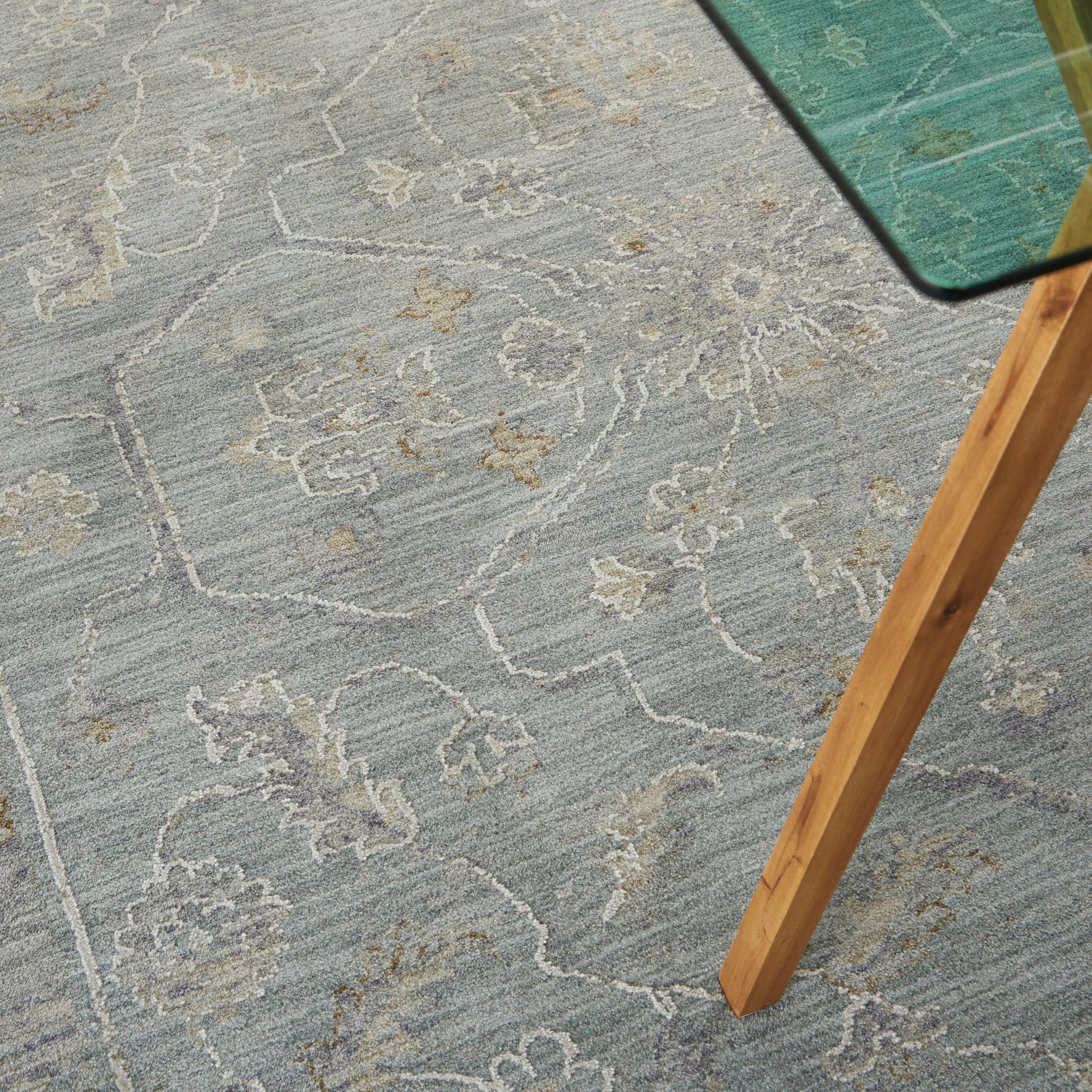 Light blue floral patterned area rug with ivory detailing; glass-topped table with a natural wood leg.