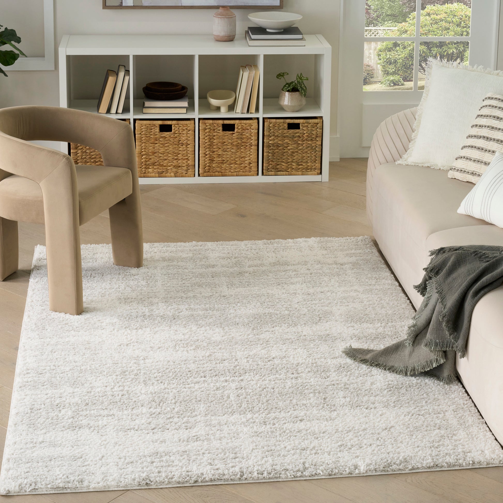 Neutral living room with beige upholstered chair, ivory rug, white cube shelf with wicker baskets, beige sofa, wood floors.