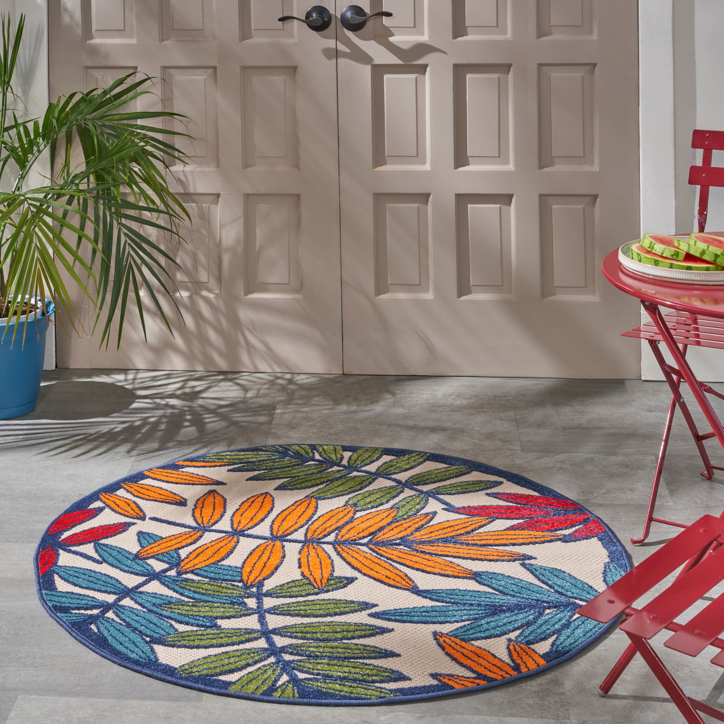 Round outdoor rug with colorful palm leaf pattern in blue, orange and green on a concrete patio beside red metal chairs and a potted plant.