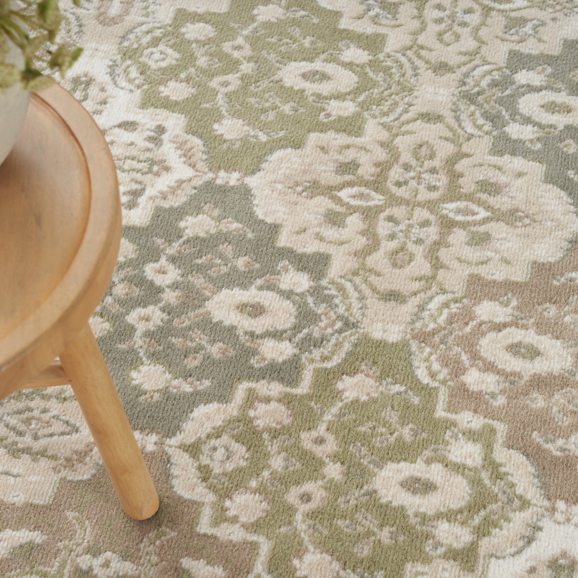 Muted olive-green and cream ornate floral area rug with vintage-style pattern; wooden stool leg visible.