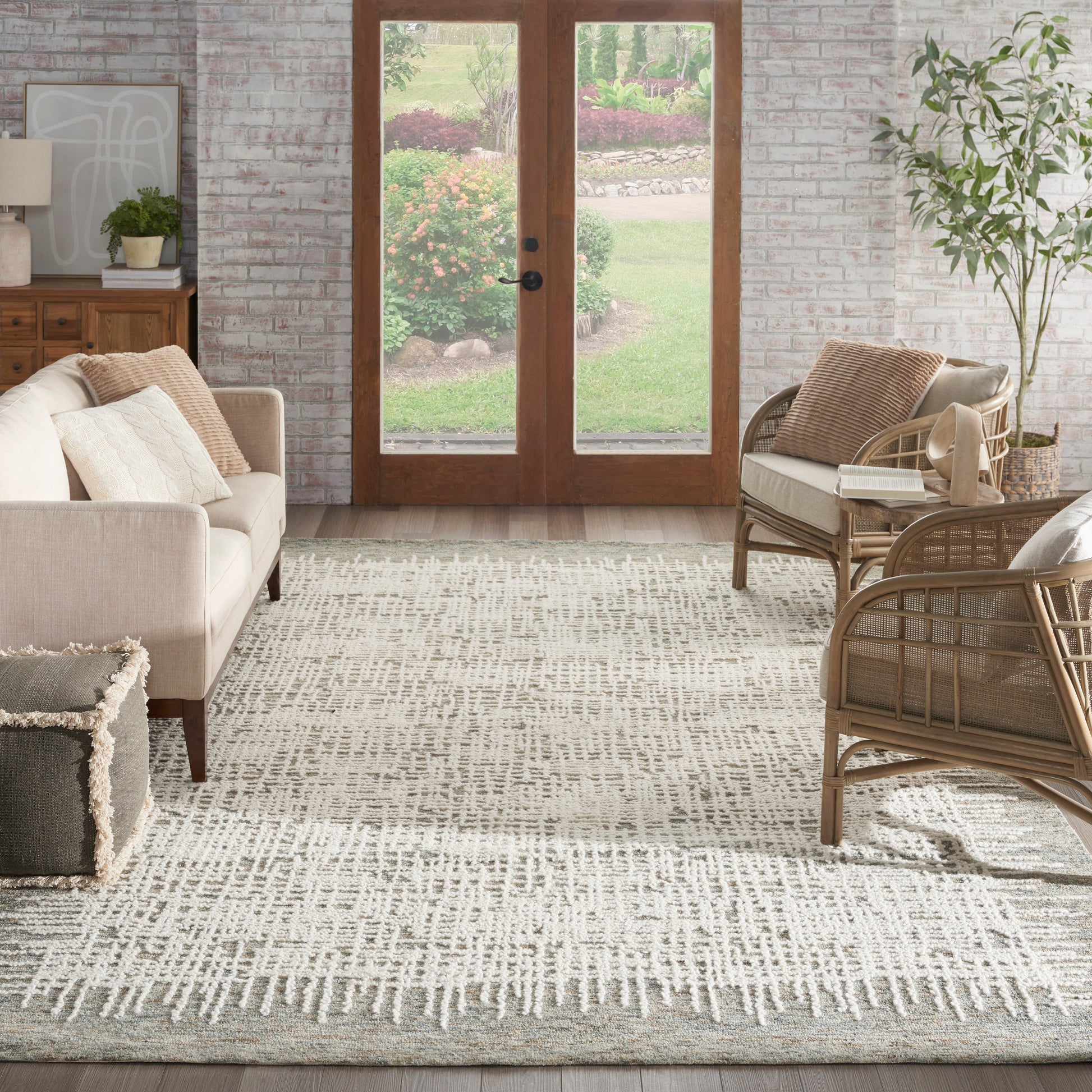 Neutral-toned living room with a textured cream-gray rug, beige sofa, rattan chairs, brick walls, and wood-framed glass doors to the garden.