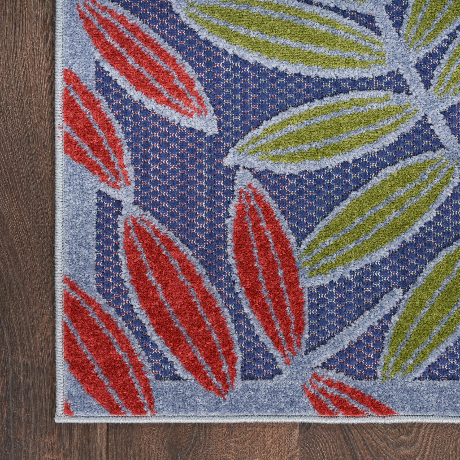 Blue woven outdoor rug with red and lime leaf patterns and a light gray border on an outdoor surface.