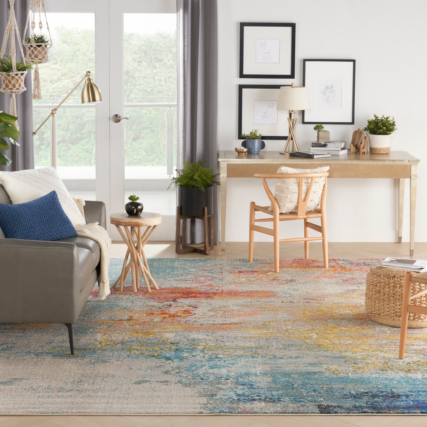 Bright living room with gray sofa, blue cushions, wooden desk and chair, colorful abstract rug, plants, and framed art