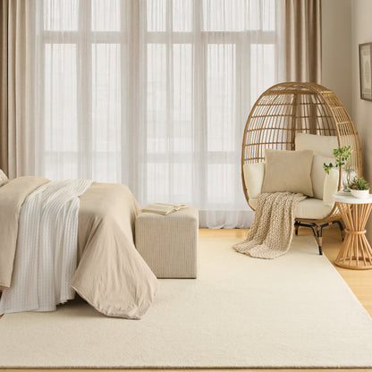 Neutral bedroom with beige bedding, cream rug, sheer curtains, and a woven rattan hanging chair with plant.