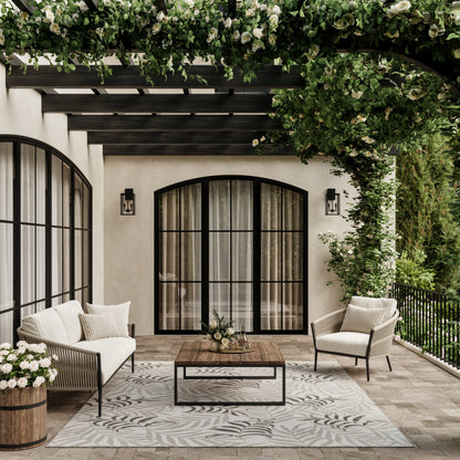 Cream patterned tropical outdoor rug on a wooden deck under a leafy pergola, with beige stucco walls and black-framed arched doors; wooden coffee table visible.