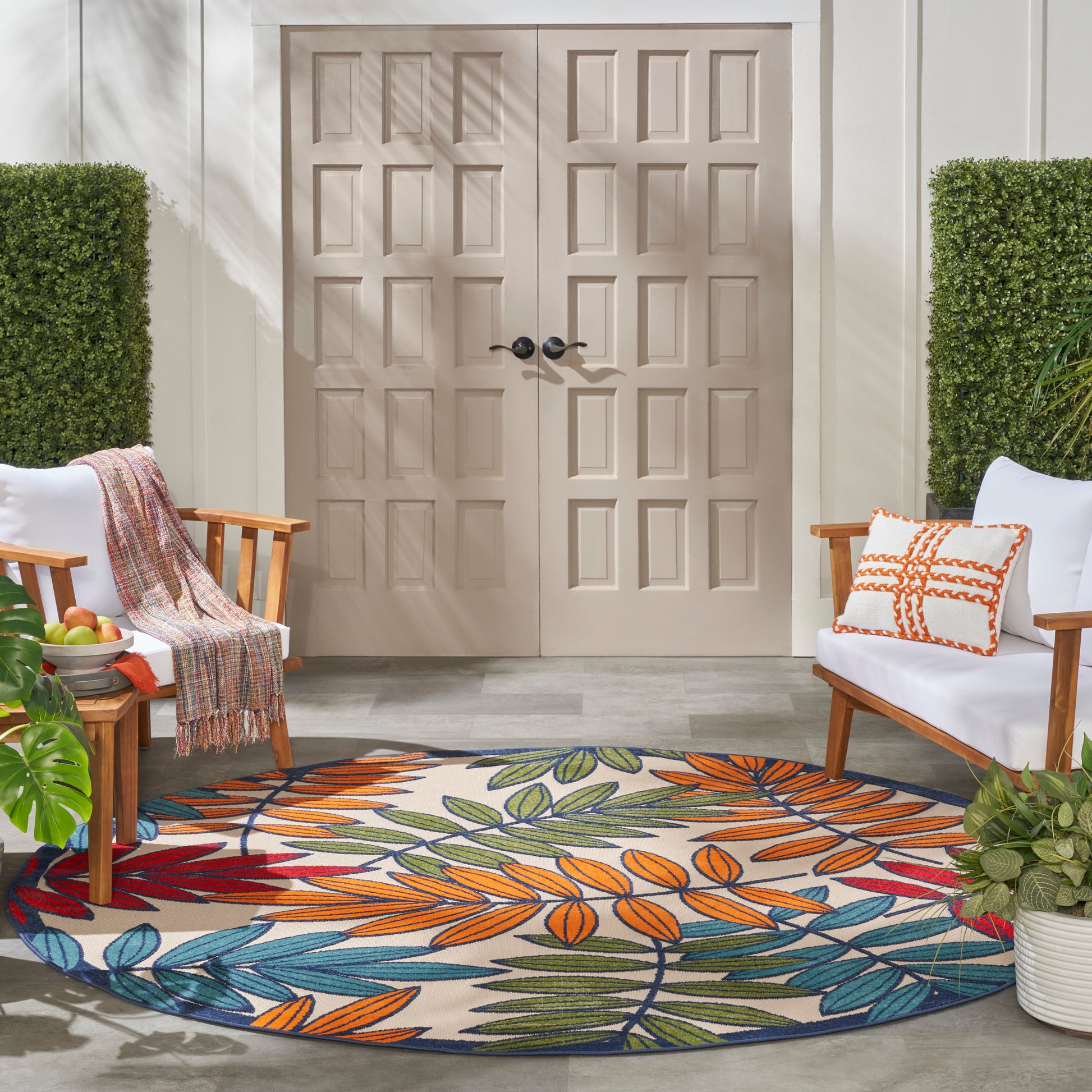 Round outdoor rug with multicolor leaf pattern on beige; wooden chairs, throw blanket, and plants on a porch.