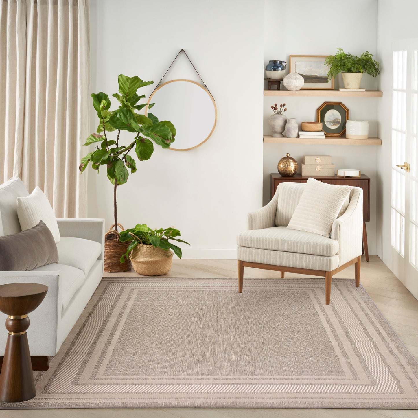 Neutral living room with beige sofa, striped armchair, woven rug, large potted plant, circular wall hoop & wood shelves with decor.