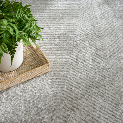 Green fern in white pot on woven rattan tray atop textured grey rug with a wave pattern.