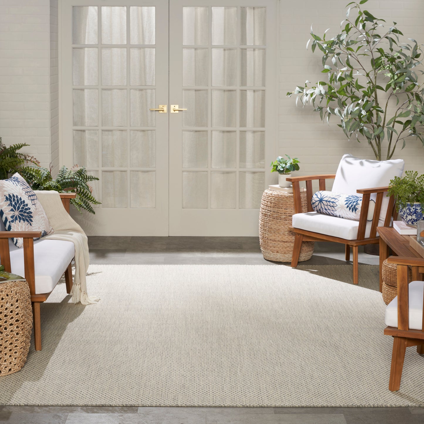 Bright sunroom with white French doors, cream rug, wooden lounge chairs with white cushions and blue pillows, surrounded by plants.