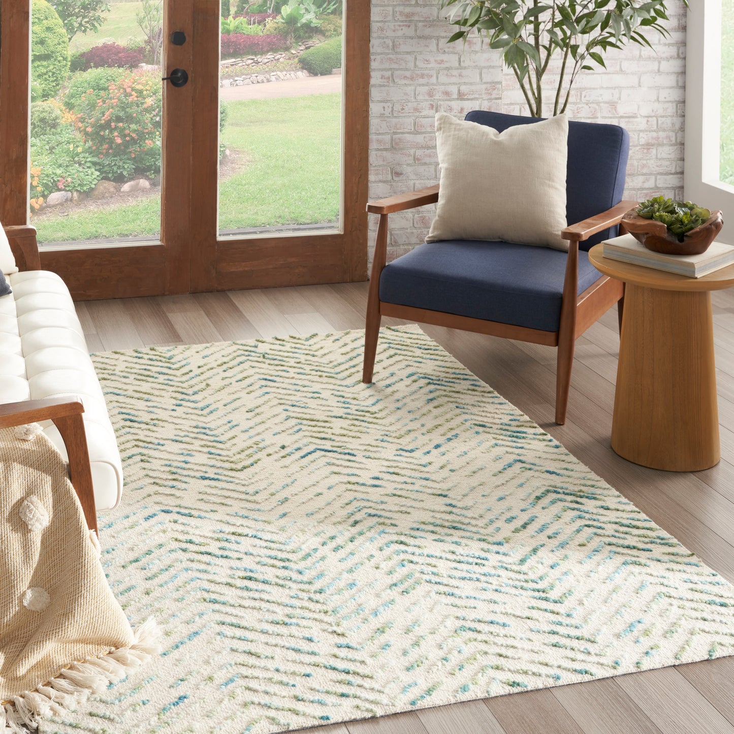 Cozy living room with cream rug and teal chevron pattern, blue armchair, round side table, brick wall, glass doors.