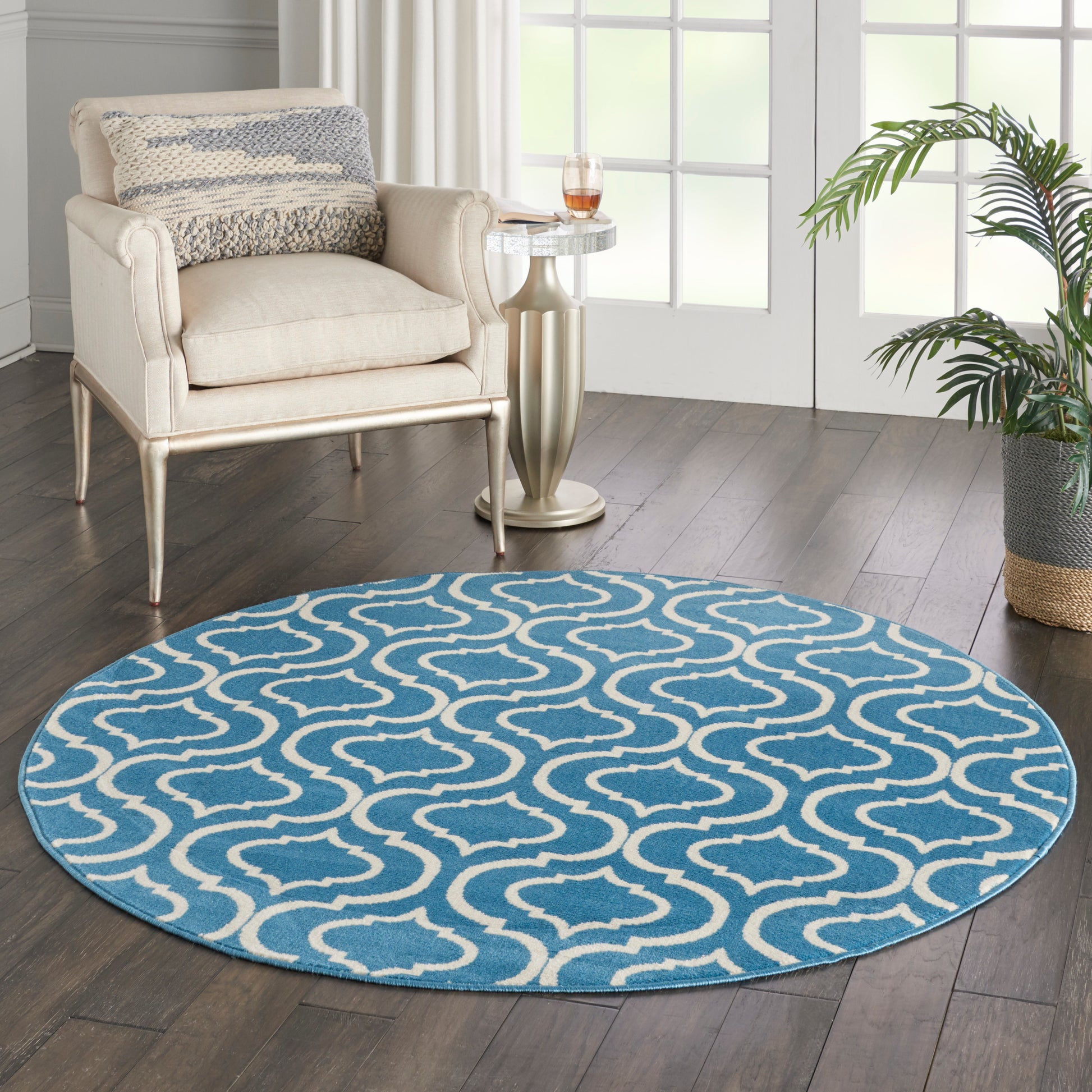 5' round blue rug with white trellis pattern on dark wood floor beside a beige upholstered armchair, glass side table, and a plant.