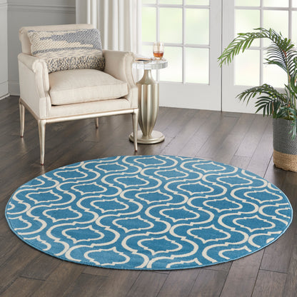5' round blue rug with white trellis pattern on dark wood floor beside a beige upholstered armchair, glass side table, and a plant.