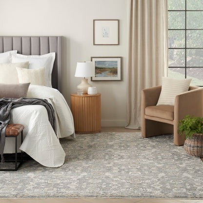 Neutral bedroom with gray tufted headboard, white quilt, tan armchair, wood nightstand, lamp, framed art, patterned rug.