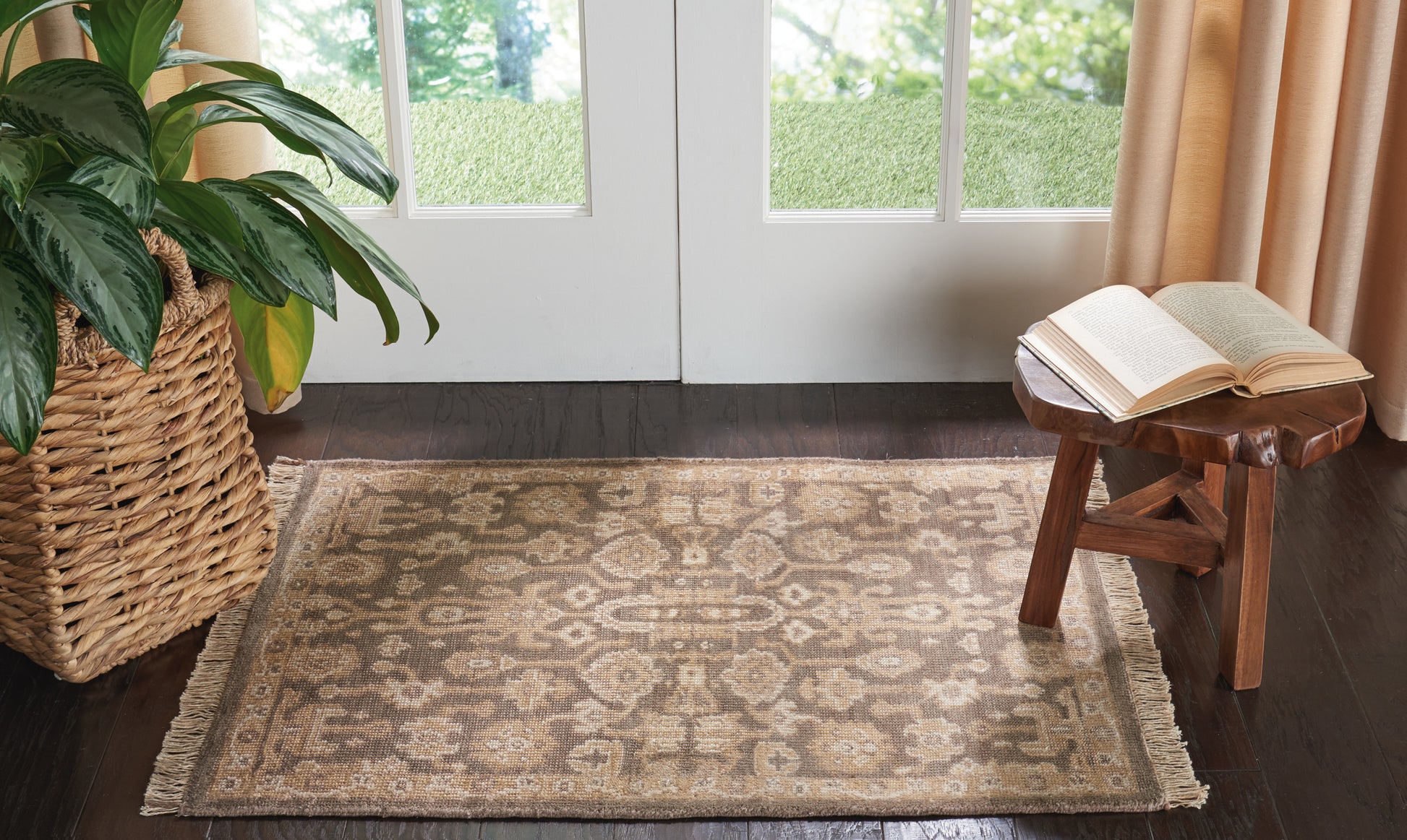 Beige patterned area rug with fringe on dark wood floor; potted plant in wicker basket left, wooden stool with book right.