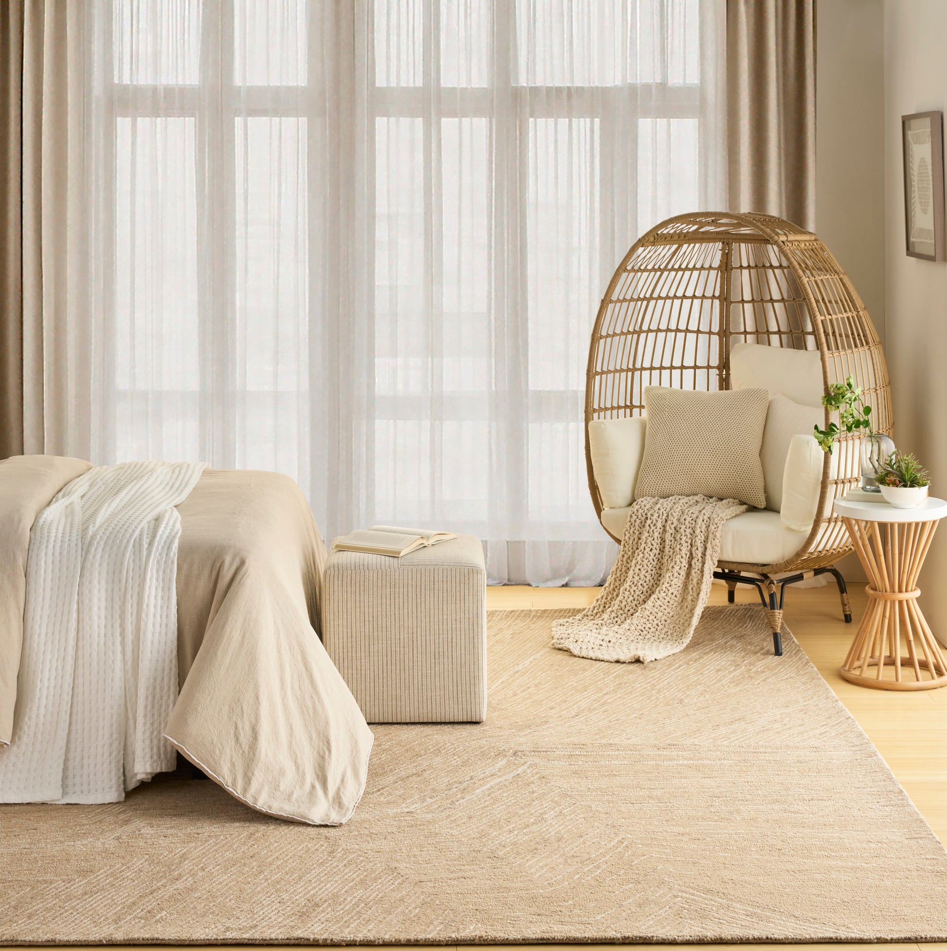 Neutral beige bedroom featuring a woven rattan egg chair, cream bedding, textured rug, and potted plant beside large sheer windows.