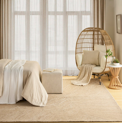 Neutral beige bedroom featuring a woven rattan egg chair, cream bedding, textured rug, and potted plant beside large sheer windows.