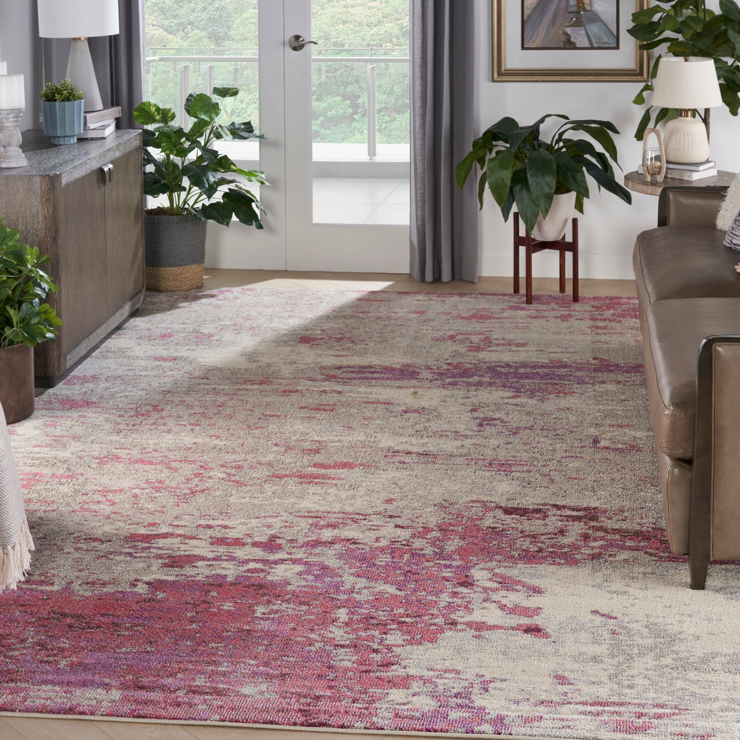 Abstract pink & beige rug anchors a contemporary living room with plants, glass doors, & brown leather sofa.