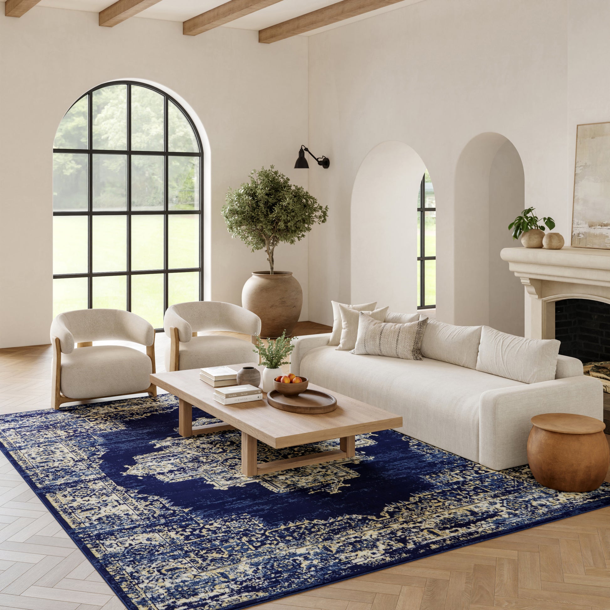 Bright living room with cream sofa, two beige armchairs, wooden beams, arched window, navy rug, coffee table, and fireplace.