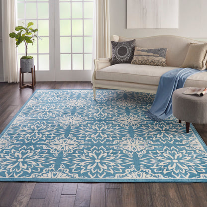 Blue and white patterned area rug anchors a cozy living room with a beige sofa, throw, ottoman, and a potted plant by French doors.