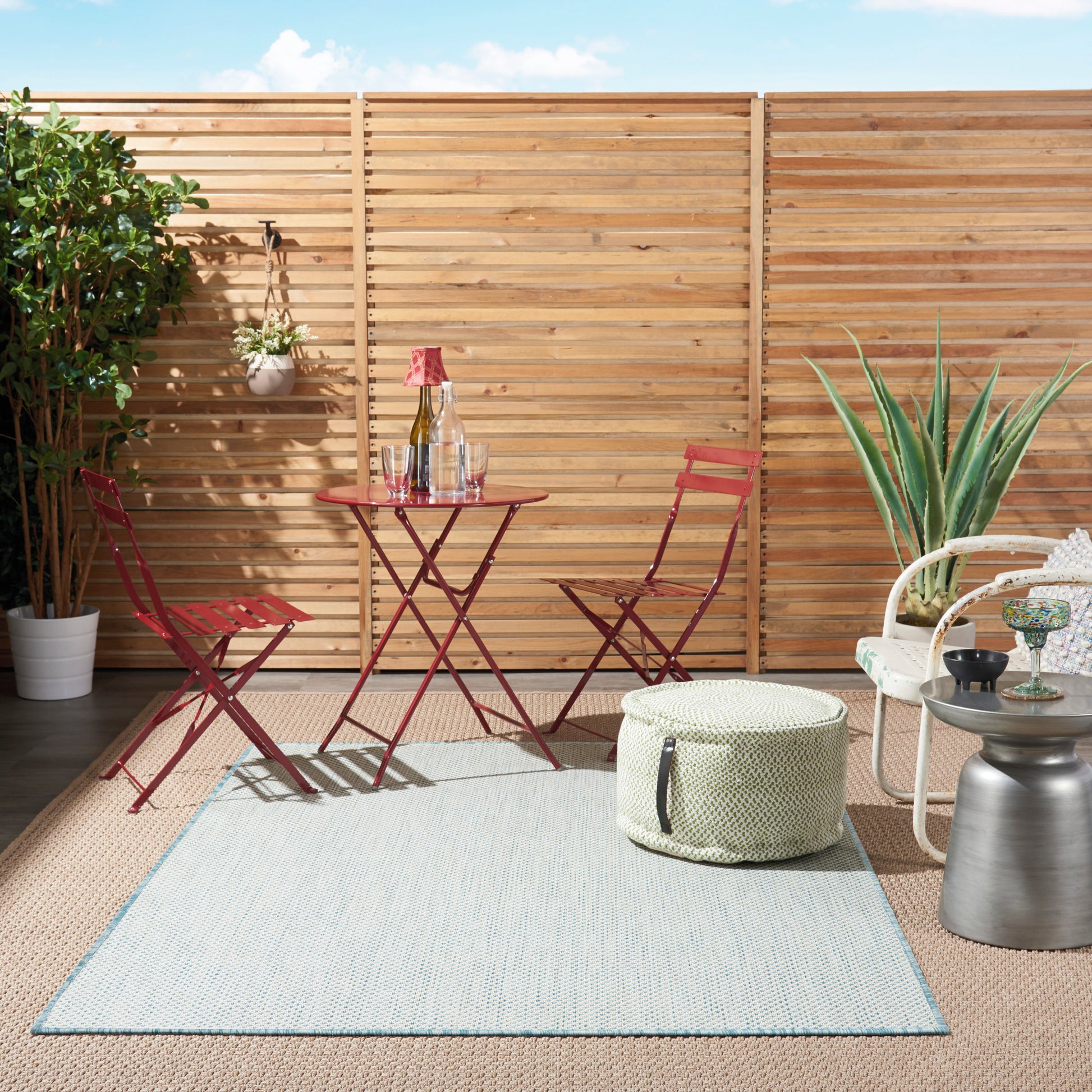 Red metal bistro set on light gray rug with blue border, wicker ottoman, white wicker chair, potted plants, wooden fence.