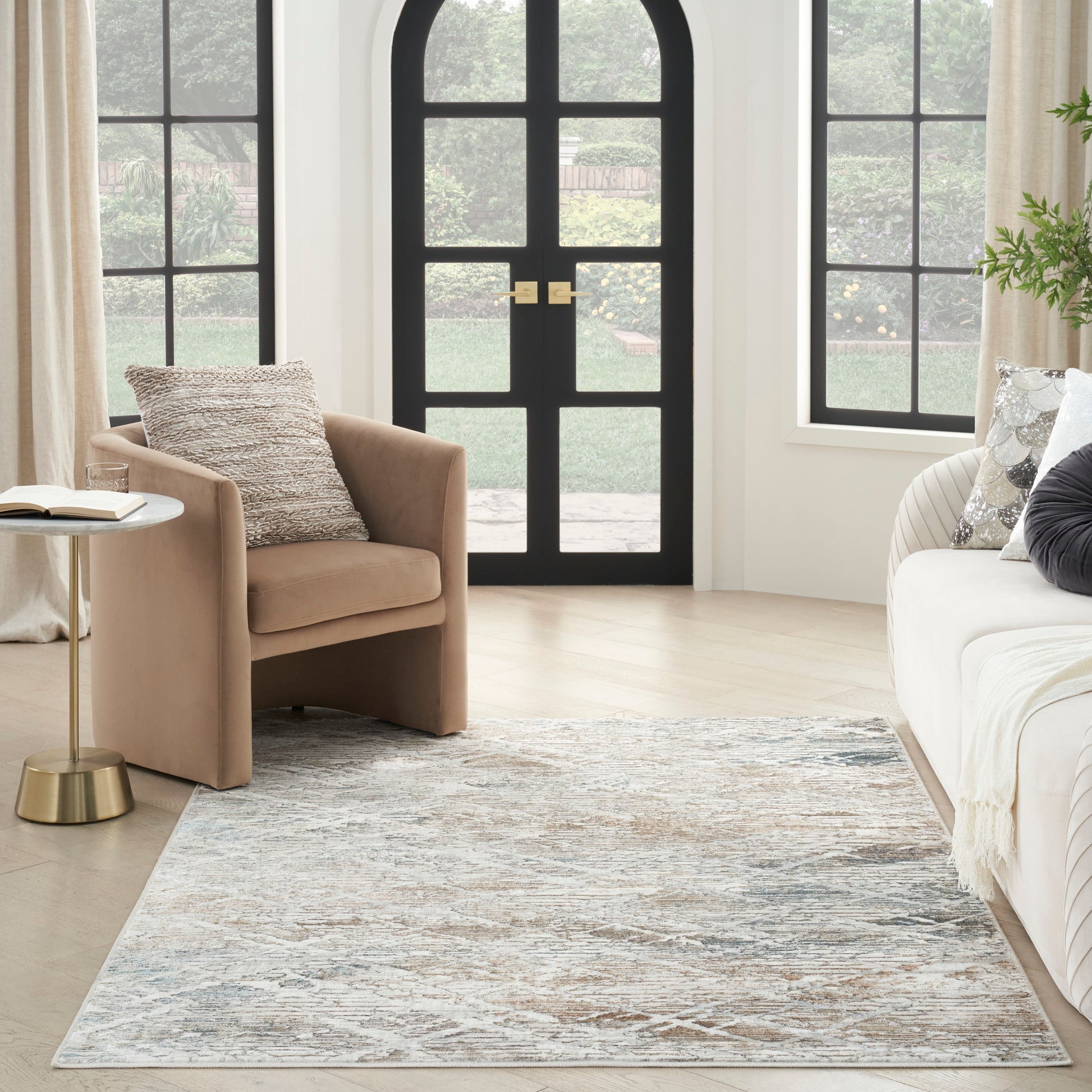 Tan armchair and cream sofa with gold side table, blue-gray abstract rug in a bright living room with arched black doors.