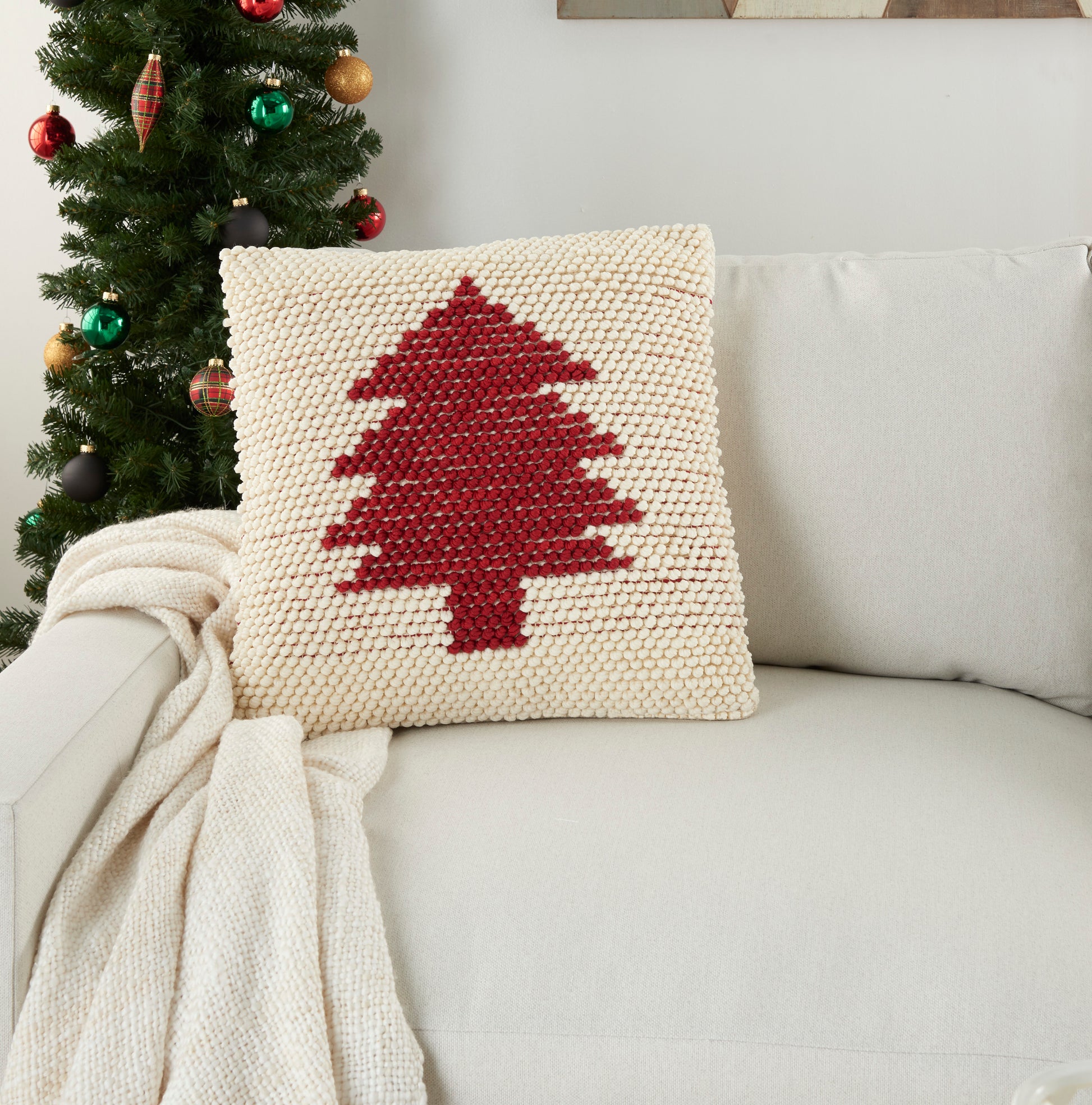Beige knitted square cushion featuring a red Christmas tree; on cream sofa with a cream throw nearby.