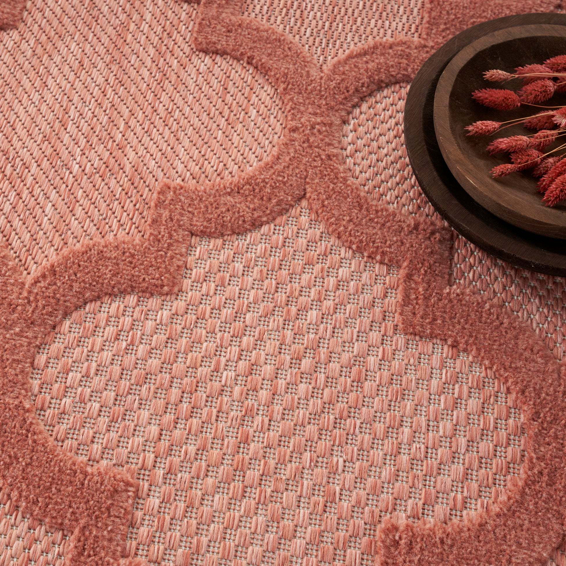 Blush pink plush rug with geometric quatrefoil pattern; dark round wooden tray with red dried botanicals in corner.