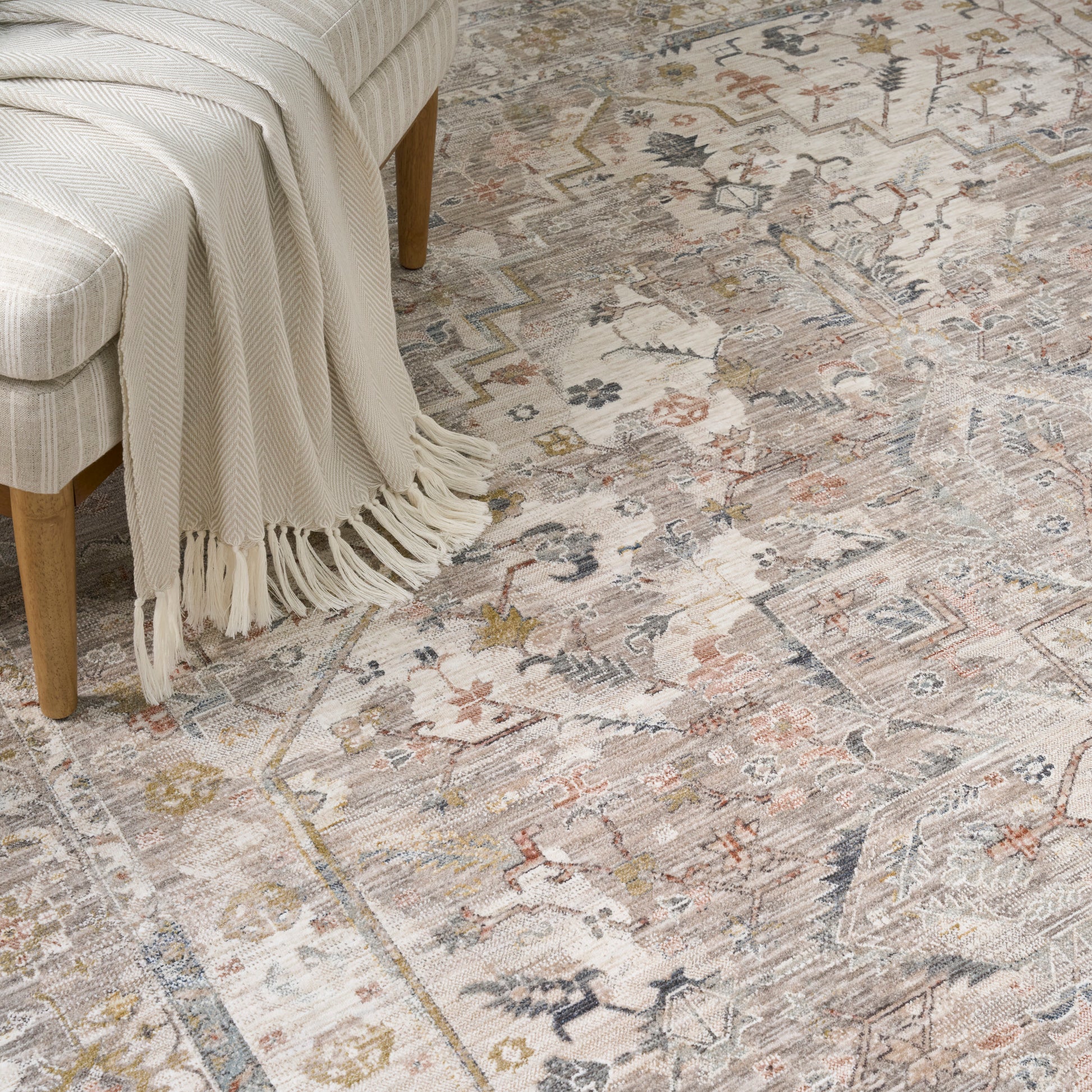 Muted floral-geometric area rug in cream, beige, blue and rust; cream knit throw draped over light wood bench.